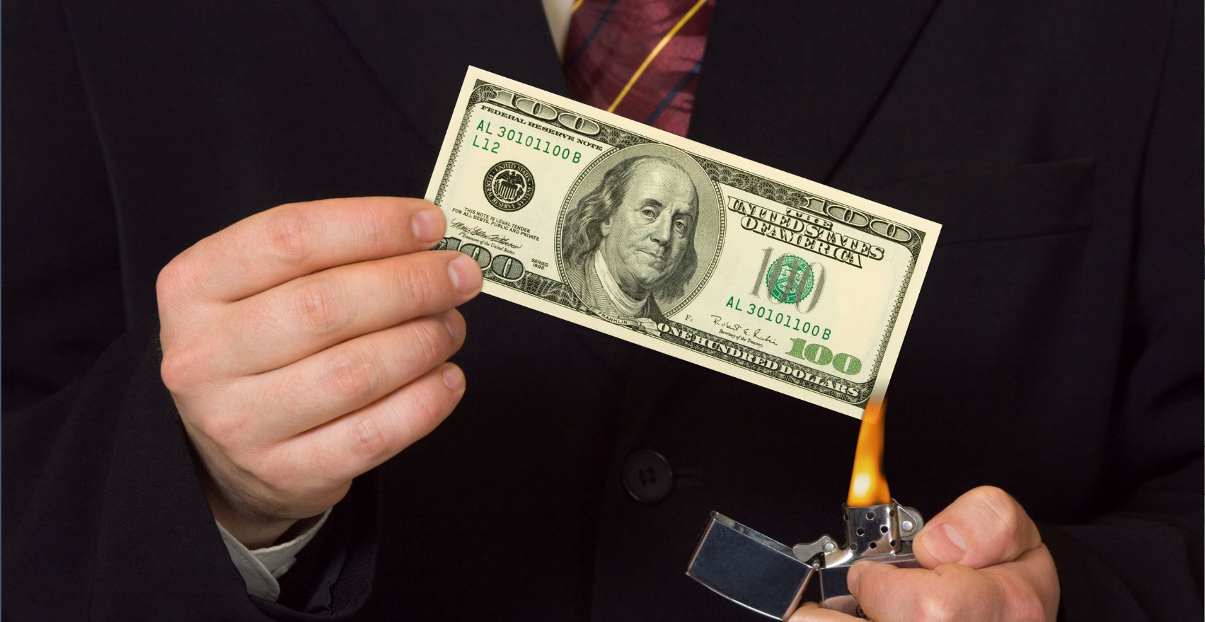 A person in a suit is holding a lighter to a $100 bill